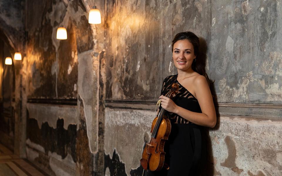 Nicola Benedetti leaning against a wall, holding her violin and smiling towards the camera