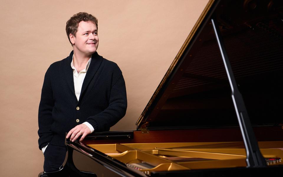 Benjamin Grosvenor standing behind a piano, looking up and to the side and smiling