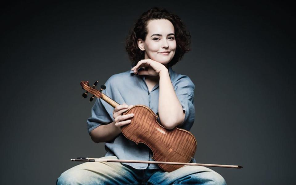 Alena Baeva sitting on Stool holding violin which is facing her. Her left arm is resting on the violin with her head resting on her hand, whilst smiling at the camera