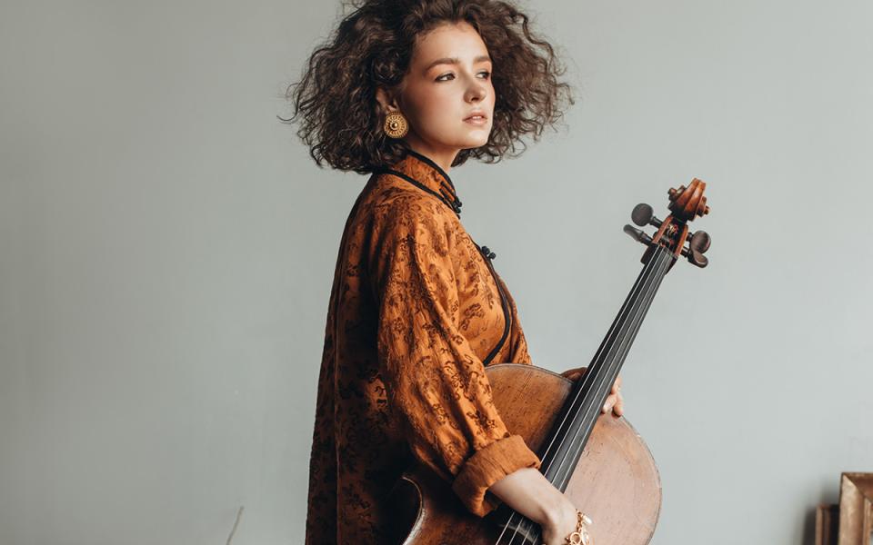 Anastasia Kobekina with her body facing the side, holding her cello. She is wearing a satin orange kimono and gold earrings