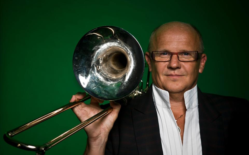 Christian Lindberg in a white shirt and black jacket, standing in front of an emerald green background. He is holding his trombone up and looking directly into the camera