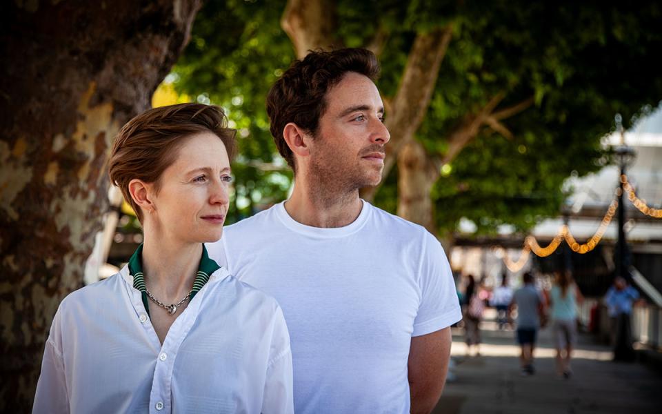 Two people stood outdoors, one in front of the other, wearing white tops, and looking towards the side